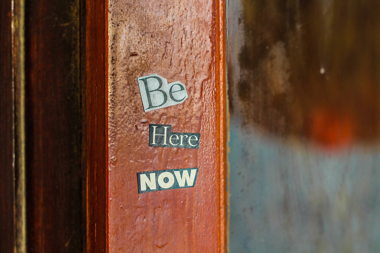 A close-up of a wooden surface with the phrase ‘Be Here Now’ in varied fonts and colors symbolizing the importance of living in the moment.